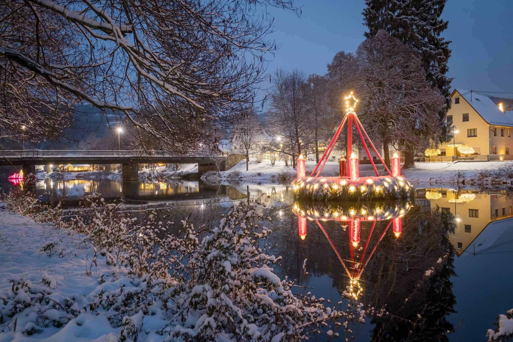 Ein großer, beleuchteter Weihnachtskranz mit Kerzen schwimmt auf einem ruhigen Fluss in einem verschneiten Dorf in der Abenddämmerung. Die Lichter spiegeln sich auf dem Wasser, und die schneebedeckten Äste und Häuser schaffen eine friedliche Winterszene.